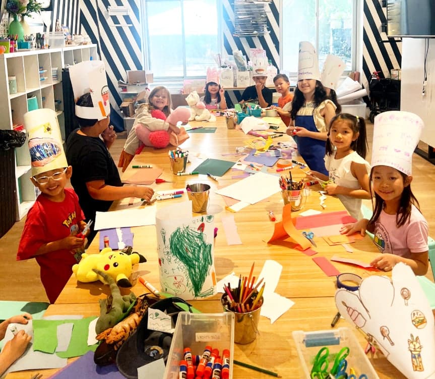 Children are happily creating art projects and decorating paper hats together at a long table in the studio, surrounded by art supplies.